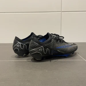 Nike Mercurial Air Zoom fotbollsskor - Snygga Nike Mercurial Air Zoom fotbollsskor i svart med blå detaljer och vita loggor. Skorna har snörning, stickad ovandel och mönstrad sula med dobbar för gräsplan. Perfekta för dig som vill ha lättviktsskor med grym känsla och respons.