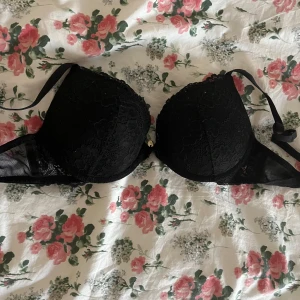 Svart spetsig bh - Snygg svart bh-topp med vadderade kupor och vacker spetsdetalj över hela framsidan. Axelbanden är justerbara och sidorna har meshpartier för extra stil. Perfekt för att ge en cool och feminin touch till din outfit.