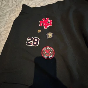 Svart kortkjol med patches från Superdry - Svart kortkjol från Superdry med coola patches och broderade märken framtill, bland annat siffran 28 och Superdry-logga. Sportig vibe och elastisk midja, perfekt för dig som gillar streetstyle och unika detaljer.