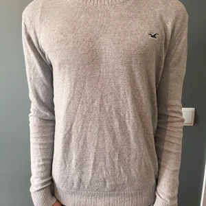 Grå stickad tröja från Hollister - Snygg grå stickad tröja från Hollister med diskret logga på bröstet. Tröjan har rund halsringning, långa ärmar och ribbade muddar. Perfekt för lager-på-lager och passar dig som är omkring 167-175. Modellen är 173.