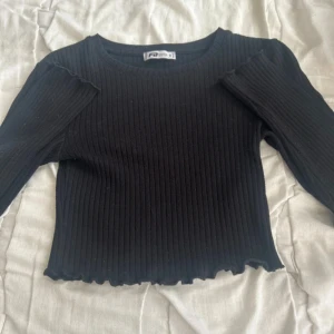 Svart ribbad långärmad topp FB Sister - Svart ribbad topp från FB Sister i storlek S. Långärmad modell med croppad passform och vågig kant nertill. Tillverkad i en mjuk mix av bomull, polyester och elastan som ger stretch och skön känsla.