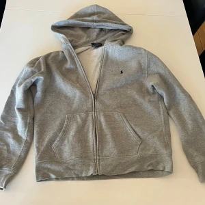 Grå ralph lauren zip hoodie - Skön ralph lauren tröja med dragkedja i grått. Den är i barnstorleken 18-20, vilket ungefär är lika stort som S/M i vuxenstorlek. Tröjan köptes för ett tag sedan, men har nästan inte använts någonting eftersom den har varit för stor för mig.