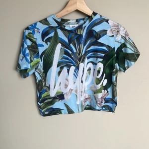 Blommig croppad topp från Hype - Säljer en croppad topp från Hype med blå botten och stora blommiga mönster i grönt, vitt och lila. T-shirten har korta ärmar och stor vit Hype-logga framtill. Den här storlek 6 men passar dig som har storlek XS