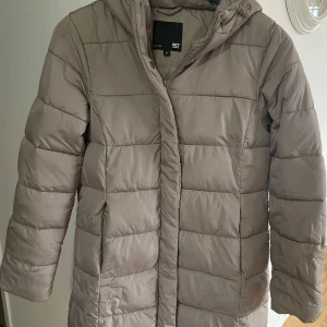 Beige pufferjacka med huva från 157 - Säljer en beige pufferjacka från 157 med huva och dragkedja. Jackan är quiltad med horisontella sömmar och har en stilren look. Perfekt för kyliga dagar och enkel att matcha med olika outfits.