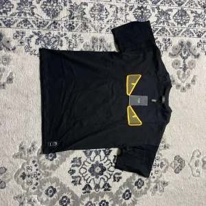 Svart Fendi t-shirt med gula detaljer - Svart t-shirt från Fendi med ikoniska gula ögon framtill. Klassisk rund halsringning och korta ärmar. Tillverkad i mjuk bomull och har en unik detalj med knappar nertill. Perfekt för dig som vill sticka ut med en lyxig streetstyle-look.