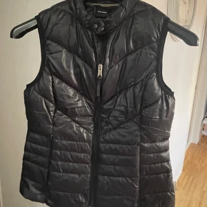 Svart dunväst från Vero Moda - Snygg svart dunväst från Vero Moda med quiltad design och dragkedja framtill. Västen har hög krage och en stilren, lätt glansig yta. Perfekt att slänga över en hoodie eller tröja för extra värme. Passar dig som gillar en clean och sportig look.