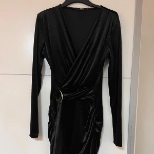 Svart sammetsklänning - Supersnygg svart sammetsklänning från Bik Bok med omlottmodell, lång ärm och markerad midja med dekorativt bälte och guldfärgad ring. Klänningen har draperad effekt och v-ringad hals.