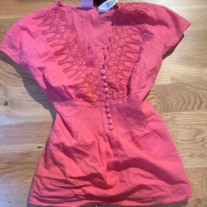 Rosa blus med spets från Emma James - Superfin rosa blus från Emma James med v-ringning, broderade spetsdetaljer och klädda knappar framtill. Knytband i midjan för snygg passform och korta ärmar. Perfekt för dig som gillar romantiska och färgstarka plagg.