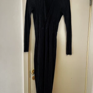 Mörklblå långklänning med omlott - Snygg mörkblå långklänning med omlott och v-ringning. Klänningen har långa ärmar och är gjord i ett plisserat tyg som ger en cool struktur. Perfekt för dig som gillar en stilren och elegant look.