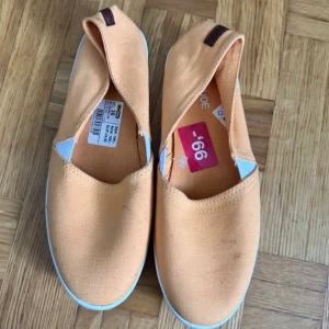 Persikofärgade slip-on sneakers - Snygga persikofärgade slip-on sneakers från KappAhl med vit sula och detaljer i ljusblått vid sidan. Skorna är i bomullstyg och har en enkel, clean look med rund tå och platt sula. Perfekta för dig som gillar en chill och stilren vibe.