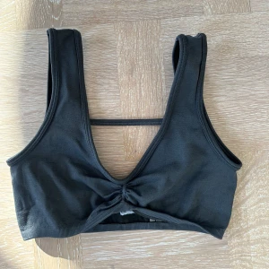 Svart croppad linnetopp med knyt - Svart croppad linnetopp med breda axelband och snygg knytdetalj framtill. Toppen har en öppen rygg med en tunn rem som ger en sportig vibe. Perfekt för att matcha med höga jeans eller shorts.