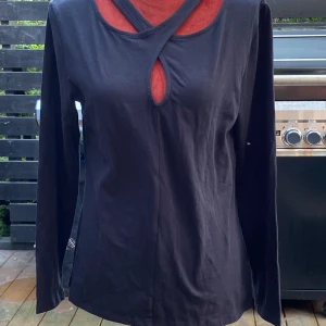 Svart långärmad topp från KappAhl L - Svart långärmad tröja med en unik halsringning som har korsade band och en liten öppning framtill. En stilren och bekväm tröja som passar bra till vardags.  L: 66 cm ﻿Bystvidd: 49 cm  Inga bilder när den sitter på mig! Katter finns i hemmet!