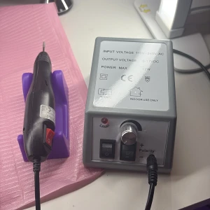 Elektrisk nagelfil med kontrollenhet - Elektrisk nagelfil med svart handtag och röd strömbrytare, tillhörande grå kontrollenhet med vred och strömbrytare. Ligger i lila hållare. Perfekt för att fixa naglarna hemma eller i salong. Drivs med sladd och har justerbar hastighet.