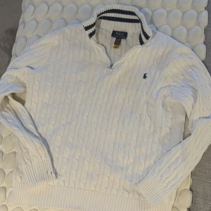Vit kabelstickad tröja Polo Ralph Lauren - Vit kabelstickad tröja från Polo Ralph Lauren i storlek XL. Tröjan har half zip, ribbade muddar och krage med marinblå ränder. Klassisk broderad logga på bröstet. Perfekt för dig som gillar preppy och stilren look.