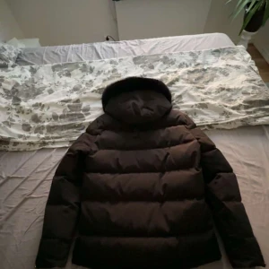 Svart dunjacka med päls från Moose Knuckles - Säljer en svart dunjacka från Moose Knuckles med fluffig päls runt huvan och snygga metalldetaljer på ärmen. Jackan har dragkedja framtill, stora fickor och en riktigt varm, puffig look. Perfekt för kalla dagar när du vill hålla stilen.