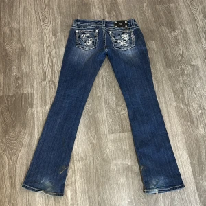 Miss Me blå bootcut jeans med detaljer - Snygga blå jeans från Miss Me i bootcut-modell med slitningar och blingade detaljer på fickorna. midja 79cm & innerben 85cm. Storlek 27 
