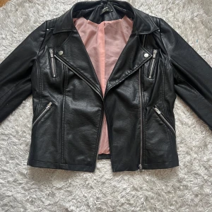Svart skinnjacka från ONLY - Snygg svart skinnjacka från ONLY med klassisk bikerstil, silverfärgade dragkedjor och detaljer. Jackan har lång ärm, flera fickor med dragkedja och en ljusrosa insida. Endast använd en gång då jag fick den som present men inte är i min stil. Originalpris 699kr men säljer den för 200kr.