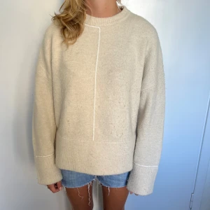 Beige stickad tröja från Zara - Säljer en oversized beige stickad tröja från Zara med lång ärm och ribbade muddar. Tröjan har en clean look med en vit söm som går vertikalt både fram och bak. Perfekt för dig som gillar en chill och enkel stil.