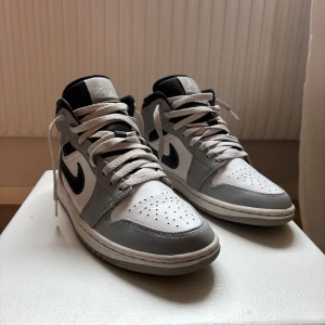 Nike Air Jordan 1 Mid vit/grå/svart - Nike Air Jordan 1 Mid sneakers i vitt, grått och svart. Klassisk siluett med svart swoosh, perforerad tå och snörning. Ovandel i skinn och syntet, svart vadderad plös och krage. Ikonisk Jordan Wings-logga på sidan. Perfekta för dig som gillar streetstyle.