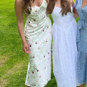 Vit blommig långklänning med snörning - Superfin långklänning i vit satin med blommigt mönster i rosa, rött och grönt. Klänningen har smala axelband, v-ringning med snörning framtill och en figurnära passform. Perfekt för sommarens alla tillfällen och ger en romantisk vibe.