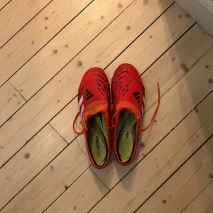 Adidas Predator elite röda fotbollsskor - Snygga Adidas Predator fotbollsskor i rött med svarta och vita detaljer. Skorna har snörning, mönstrad ovandel och dobbar för bra grepp på planen. Perfekta för dig som vill sticka ut på matchen med stil och funktion.