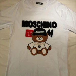 Vit Moschino t-shirt med nalletryck - Snygg vit t-shirt från Moschino med ett stort broderat nallemotiv framtill och tydlig Moschino-logga. T-shirten har rund hals, korta ärmar och är gjord i mjuk bomull. Perfekt statement-plagg för dig som gillar lekfull design och streetstyle. Storlek medium, knappt använd 