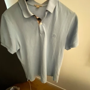 Ljusblå pikétröja från Burberry Brit - Snygg ljusblå pikétröja från Burberry Brit med klassisk krage och korta ärmar. Tröjan har diskret broderad logga på bröstet och ikoniskt rutigt tyg vid knäppningen. Tillverkad i mjuk bomull som känns skön mot huden.