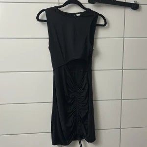 Svart kort klänning med snörning och öppen vid magen - Snygg svart kort klänning med ärmlös design och cool cut-out framtill. Klänningen har trendig snörning och rynkdetalj längs magen som ger en unik look. Perfekt för dig som gillar stilrena och moderna plagg.