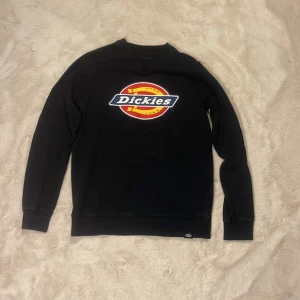 Svart Dickies sweatshirt  - Svart sweatshirt från Dickies i storlek small. Tröjan har en stor färgglad Dickies-logga på bröstet och är långärmad med ribbade muddar. Perfekt för dig som gillar streetwear och vill ha en enkel men snygg look.