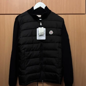 Svart Moncler cardigan med stickade ärmar - Svart cardigan från Moncler med quiltad front och stickade ärmar. Jackan har dragkedja, två fickor framtill och Moncler-logga på bröstet. Först till kvarn!🙏✌️NFC scan funkar.