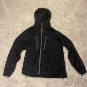 Svart regnjacka från True North - En svart stilren sportig regnjacka ifrån True North, perfekt för dig som vill springa när det regnar ute. Två fickor på bröstet, två fickor på sidan och två innerfickor. I storlek S 