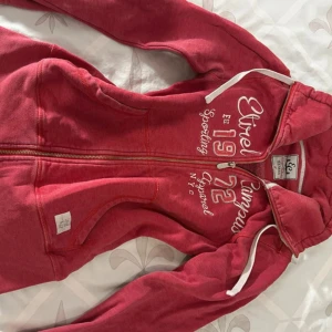 Rosa zip-up hoodie från Etirel - Röd hoodie från Etirel med vit text och brodyr på bröstet. Tröjan har huva med vita snören, hel dragkedja och två fickor framtill. Perfekt för chill dagar eller när du vill ha en sportig look. Mjuk bomullskänsla och långärmad modell.