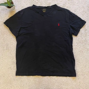 Svart v-ringad t-shirt från Ralph Lauren - Säljer en klassisk svart v-ringad t-shirt från Ralph Lauren med den ikoniska röda loggan broderad på bröstet. Perfekt för en stilren och avslappnad look. Tillverkad i mjuk bomull för extra komfort och i perfekt skick