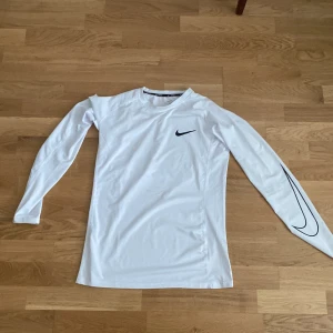 Vit långärmad Nike Pro träningströja - Nike Pro långärmad träningströja i vitt med svart swoosh-logga på bröstet och ärmen. Tight passform och tillverkad i ett lätt, ventilerande syntetmaterial som andas. Perfekt för intensiva träningspass eller sportaktiviteter. aldrig använt, storlek L fast skulle kunna passa M.
