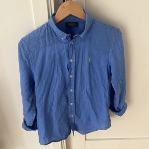 Blå linneskjorta från Polo Ralph Lauren - Snygg blå skjorta från Polo Ralph Lauren i luftig linne. Klassisk krage, knäppning framtill och ikoniska gula broderade loggan på bröstet. Perfekt för dig som gillar stilren och avslappnad look. Passar dig som är ca 165. 