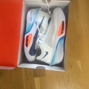 Nike storlek 43 next 3  löpar skor med kvitto - Säljer ett par riktigt snygga Nike sneakers i vitt med blå swoosh och coola orange detaljer på sulan. Ovandelen är i stickad mesh för maxad andningsförmåga och komfort. Sulan har en gradient i blått och vitt, perfekt för dig som gillar sportig och modern stil.