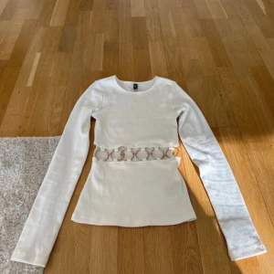 Vit långärmad topp med cut out från Divided - Vit långärmad topp från Divided i bomullsmix. Snygg cut out-detalj med ringar i midjan som ger en unik look. Tight passform och rund halsringning. Perfekt för dig som vill sticka ut lite extra. Aldrig använd