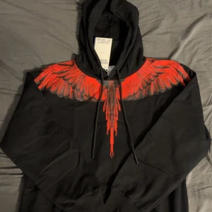 Svart hoodie Marcelo Burlon med röda vingar - Svart hoodie från Marcelo Burlon County of Milan med ikoniskt rött vingtryck över axlar och bröst. Tröjan har huva med snörning och långa ärmar. Grym streetwear-vibe och riktigt snygg detalj med vingarna som poppar mot det svarta tyget.