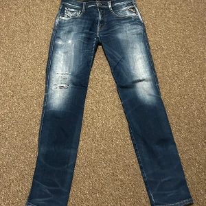 Blå Replay jeans med slitningar - Snygga blå jeans från Replay med slitna detaljer och ljusa partier framtill. Modellen är stretch och klassisk femficksmodell med raka ben och kontrastsömmar. Märkespatch bak i midjan. Hål i byxan även baktil vid rumpan så det är ett minus. Kom gärna med prisförslag!!