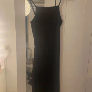 Svart långklänning med smala axelband - En stilren svart långklänning med smala axelband och rak siluett. Klänningen har minimalistisk design och passar perfekt för dig som gillar en enkel men elegant look. Perfekt att styla med smycken eller en cool jacka.