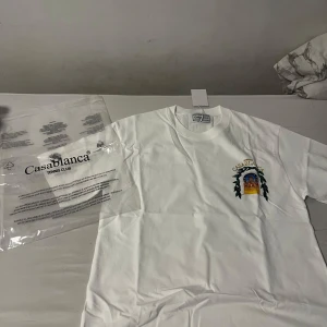 Vit t-shirt från Casablanca med tryck - Vit t-shirt från Casablanca med färgglatt tryck av en byggnad och gröna blad på ryggen samt mindre tryck på bröstet. Klassisk passform och rund hals. Perfekt för dig som vill sticka ut med en snygg och unik tee.