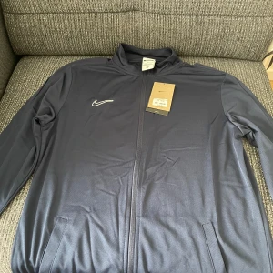 Mörkblå Nike träningsjacka Dri-Fit - Snygg mörkblå träningsjacka från Nike med Dri-Fit-teknologi och slim fit-passform. Jackan har hel dragkedja framtill, ståkrage och diskret Nike-logga på bröstet. Perfekt för dig som vill ha en sportig look och bekväm känsla under träningen.