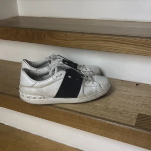 Valentino Open vita sneakers med svart detalj - Snygga vita Valentino Open sneakers med bred svart läderdetalj över sidan. Klassisk rund tå, snörning och platt sula. Skorna är i skinn och har en stilren, modern look som passar till det mesta. Perfekta för dig som gillar exklusiva märken och streetstyle.