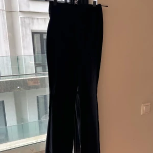 Svarta kostymbyxor från NA-KD - Snygga svarta kostymbyxor från NA-KD i klassisk tight modell med utsvängning och slits nertill. Byxorna har hög midja och raka ben, perfekta för en clean och stilren look. Materialet känns mjukt och följsamt, vilket gör dem bekväma att bära hela dagen.