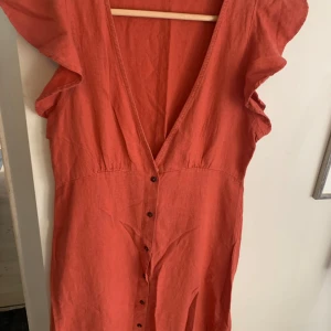 Röd långklänning från ONLY med knappar - Snygg röd långklänning från ONLY med djup v-ringning, volangärmar och knappar hela vägen framtill. Tillverkad i ett lätt och luftigt linnematerial, perfekt för varma dagar. Klänningen har en avslappnad passform och är superenkel att styla.