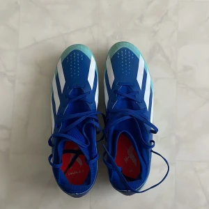 Adidas X Crazyfast.1. - Snygga blå fotbollsskor från adidas med vita ränder och ljusblå detaljer. Skorna har snörning och en sula med dobbar för bra grepp på planen. Ovandelen är i syntetmaterial och insidan har en röd innersula med adidas-logga. Perfekta för dig som vill sticka ut på matchen.