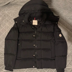 Svart Moncler pufferjacka med huva - Säljer en svart pufferjacka från Moncler med avtagbar huva och klassisk logga på ärmen. Jackan har dragkedja och knappar framtill, samt flera fickor. Insidan har ett unikt serietryck. Perfekt för kalla dagar och riktigt snygg streetstil. Strlk 2=M