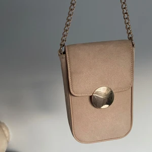 Beige axelväska med guldkedja - Snygg beige axelväska i mocka med guldfärgad kedja och rund metallknapp framtill. Perfekt storlek för mobilen och det viktigaste. Stilren och enkel design som passar till många outfits.