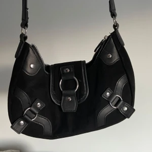 Svart axelväska med metalldetaljer - Cool svart axelväska med snygga metalldetaljer och ringar framtill. Väskan har dragkedja upptill och dekorativa remmar i skinnimitation. 