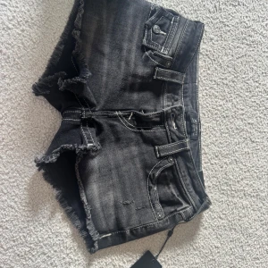 Svarta jeansshorts True Religion - Snygga svarta jeansshorts från True Religion med låg midja och råa, fransiga kanter. Shortsen har tydliga vita kontrastsömmar, klassiska fickor med lock och knappar bak samt bälteshällor. Perfekta för en edgy och avslappnad stil.